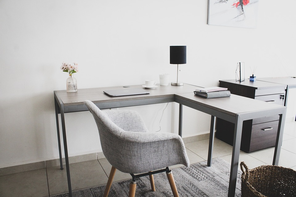 home office with a laptop, small lamp with a black shade, and binder sitting on an L shaped desk with a gray chair