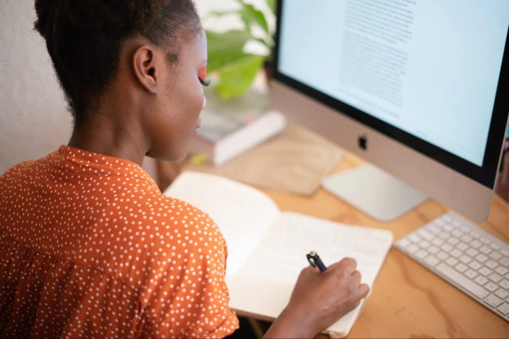 woman is taking notes in a notebook at a desk with a computer